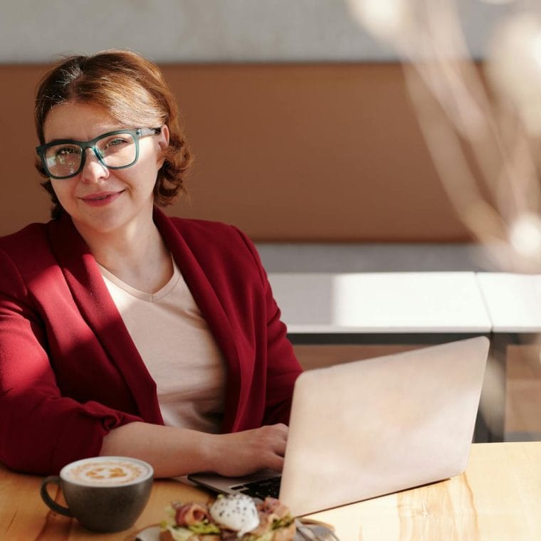 Smiling woman in red blazer using a laptop at a café table, enjoying her workday