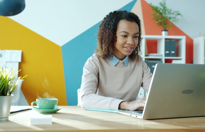 A smiling woman works on a laptop at a desk.